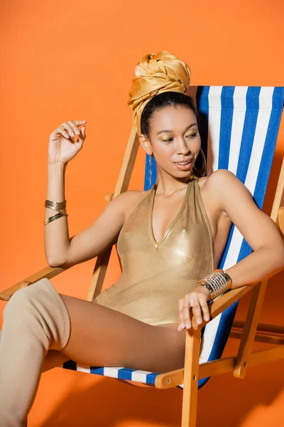 Trendiges afrikanisch-amerikanisches Model im Badeanzug lächelt, während es auf einem Liegestuhl auf orangefarbenem Hintergrund sitzt — Stockfoto