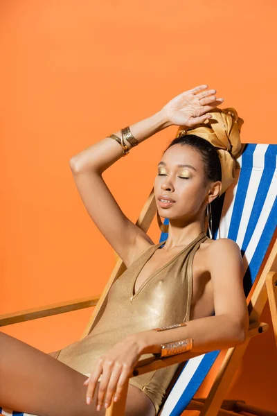 Modelo americano africano elegante em maiô e faixa de cabeça sentada na cadeira de praia em fundo laranja — Fotografia de Stock