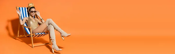 Mulher americana africana na moda e sorridente em roupa de banho acenando a mão enquanto sentada na cadeira de praia em fundo laranja, banner — Fotografia de Stock