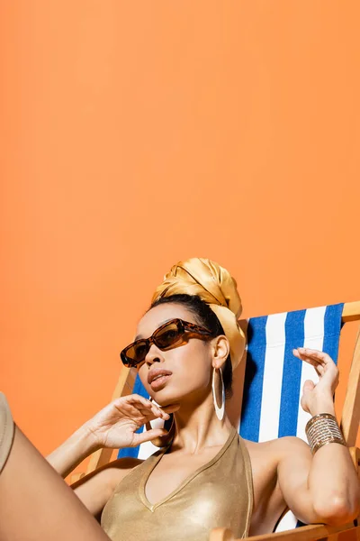 Fashionable african american model in swimsuit and headscarf sitting on deck chair isolated on orange — Stock Photo