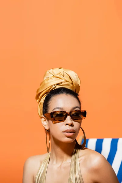 Retrato do modelo americano africano na moda em lenço de cabeça e óculos de sol olhando para longe perto da cadeira de praia isolada em laranja — Fotografia de Stock