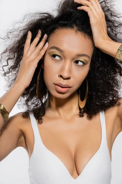 Joven modelo afroamericano ajustando el pelo rizado mientras mira hacia otro lado aislado en gris - foto de stock