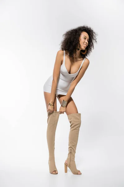 Full length of curly african american woman in mini dress adjusting knee boot on white — Stock Photo