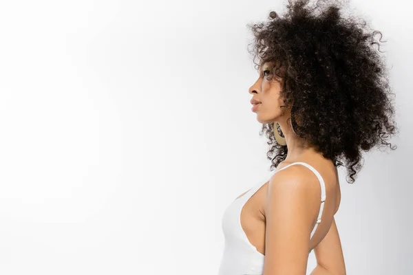 Vue latérale du jeune modèle afro-américain aux cheveux bouclés posant en maillot de bain blanc sur fond gris — Photo de stock