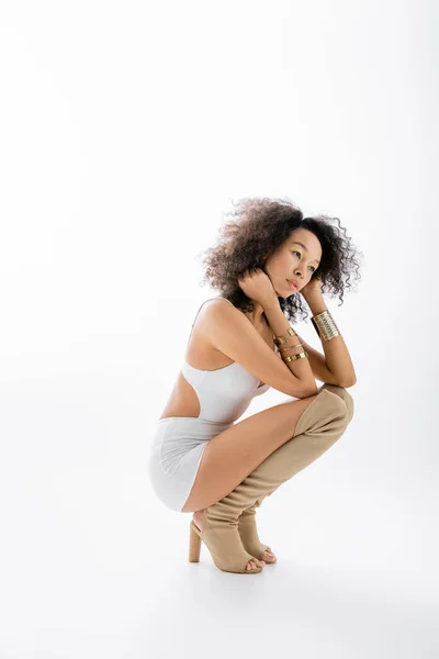 Toute la longueur de joli modèle afro-américain en mini robe et bottes de genou assis sur fond gris — Photo de stock
