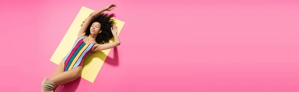 Vue du haut du modèle afro-américain bouclé en maillot de bain rayé coloré couché sur fond jaune et rose, bannière — Photo de stock