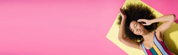 Вид зверху насолодженої афроамериканської моделі в барвистому смугастому купальнику, лежить на жовтому і рожевому фоні, банер — стокове фото