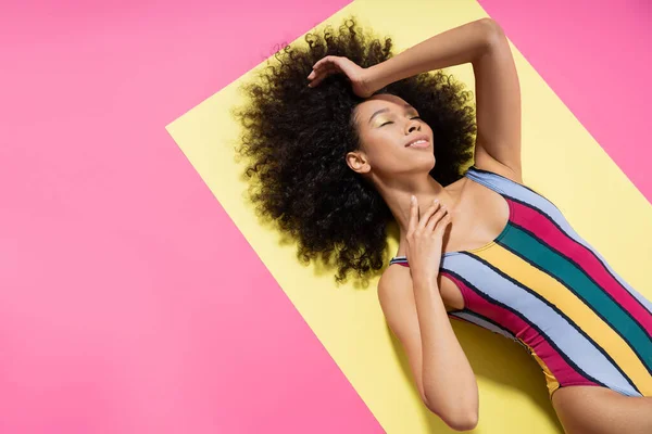 Top view of satisfied african american model in colorful striped swimsuit lying on yellow and pink background — Stock Photo