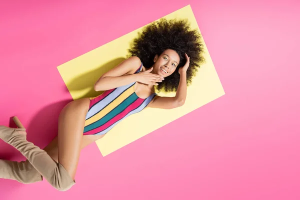 Вид зверху весела афро-американська модель в смугастому купальнику і довгі чоботи лежать на жовтому і рожевому тлі — стокове фото