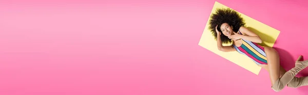 Вид зверху щасливої афроамериканки в смугастому купальнику і довгі чоботи лежать на жовтому і рожевому фоні, банер — стокове фото