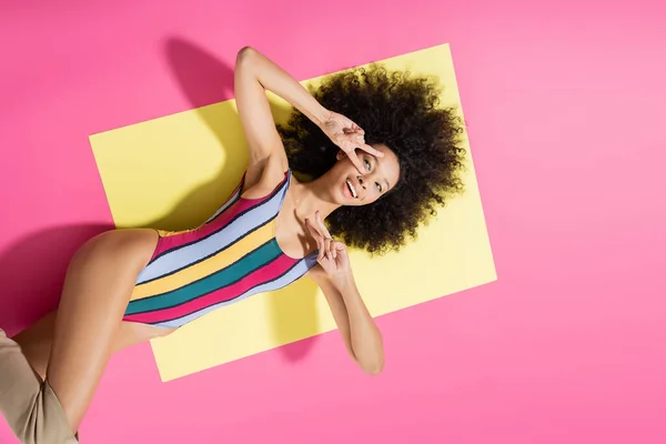Vista superior del alegre modelo afroamericano en traje de baño a rayas mostrando signo v mientras está acostado sobre fondo amarillo y rosa - foto de stock