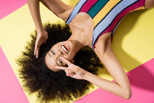 Vue du dessus du modèle afro-américain excité en maillot de bain rayé montrant le signe v tout en étant couché sur fond jaune et rose — Photo de stock
