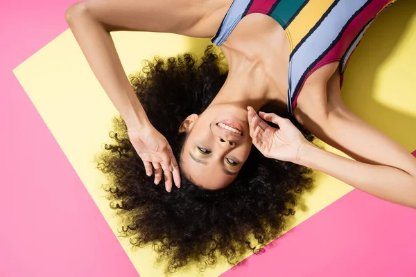 Top view of smiling african american woman in striped swimsuit lying on yellow and pink background — Stock Photo