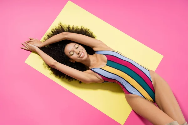 Top view of pleased african american woman in colorful swimsuit getting tan on yellow and pink background — Stock Photo