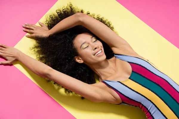 Top view of happy african american woman in colorful swimsuit getting tan on yellow and pink background — Stock Photo