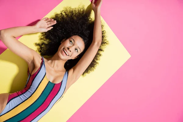 Draufsicht auf freudige afrikanisch-amerikanische Frau in bunten Badeanzug bekommen Bräune auf gelbem und rosa Hintergrund — Stockfoto
