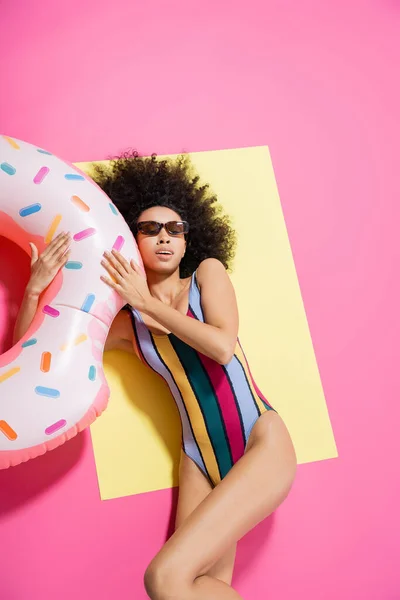 Вид зверху красивої афроамериканської жінки в купальнику і сонцезахисних окулярах, що надуваються біля надувного кільця на жовтому і рожевому — стокове фото