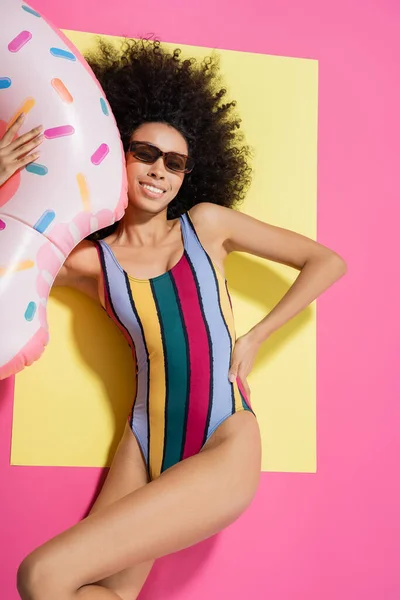 Top view of happy african american woman in swimsuit and sunglasses posing near inflatable ring on yellow and pink — Stock Photo