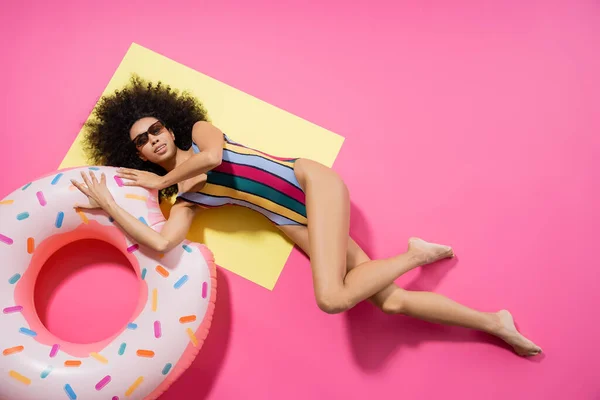 Vista dall'alto della donna afroamericana a piedi nudi in costume da bagno e occhiali da sole in posa vicino anello gonfiabile su giallo e rosa — Foto stock