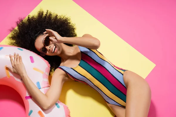 Top view of overjoyed african american model in swimsuit and sunglasses posing near inflatable ring on yellow and pink — Stock Photo