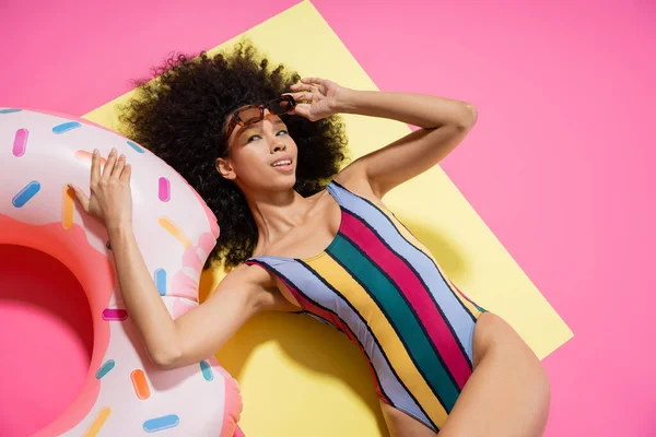 Vista superior del encantador modelo afroamericano en traje de baño y gafas de sol posando cerca del anillo inflable en amarillo y rosa — Stock Photo