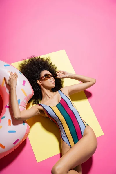 Вид сверху на очаровательную африканскую модель в купальнике и солнцезащитных очках, загорающую возле надувного кольца на желтом и розовом — стоковое фото