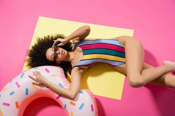Vista dall'alto del modello afroamericano in costume da bagno che regola gli occhiali da sole e si abbronza vicino all'anello gonfiabile su giallo e rosa — Foto stock