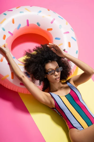 Vista dall'alto di affascinante modello africano americano in costumi da bagno a righe e occhiali da sole abbronzarsi vicino anello gonfiabile su giallo e rosa — Foto stock