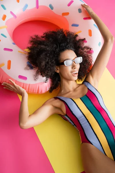 Vista superior del modelo afro-americano positivo en traje de baño a rayas y gafas de sol bronceándose cerca del anillo inflable en amarillo y rosa - foto de stock