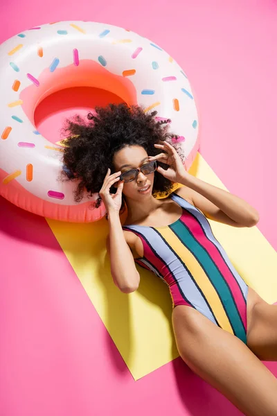 Вид зверху на афроамериканську жінку в смугастому купальнику, що регулює сонцезахисні окуляри і отримує засмагу біля надувного кільця на жовтому і рожевому — стокове фото