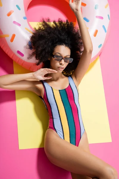 Vista superior da mulher americana africana em roupa de banho listrada e óculos de sol elegantes ficando bronzeado perto do anel inflável no amarelo e rosa — Fotografia de Stock