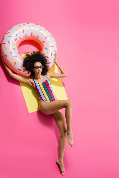 Draufsicht auf eine barfüßige Afroamerikanerin in Badebekleidung und stylischer Sonnenbrille, die sich in der Nähe eines aufblasbaren Rings in Gelb und Rosa bräunt — Stockfoto