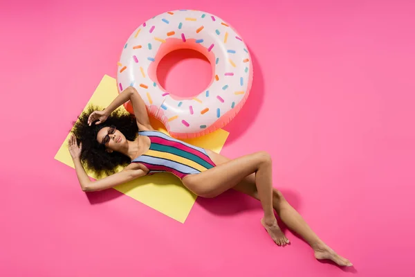 Вид зверху на афроамериканську модель босоніж у купальнику та сонцезахисних окулярах, що надуваються біля надувного кільця на жовтому та рожевому — стокове фото