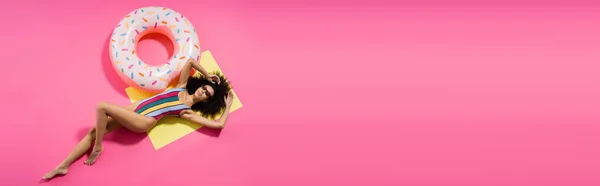Вид зверху на афроамериканську модель босоніж у купальнику та модні сонцезахисні окуляри, що надуваються біля надувного кільця на рожевому, банер — стокове фото