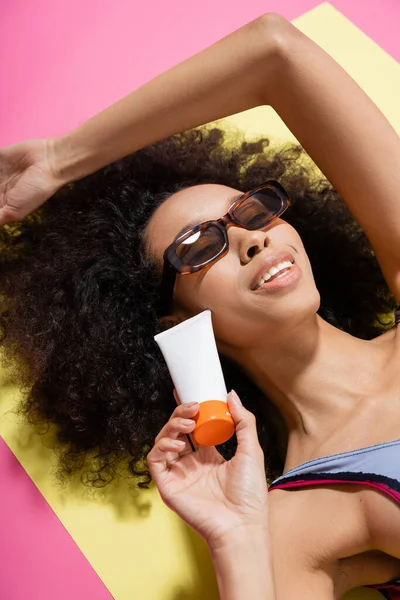Насолоджена афроамериканська жінка в модних сонцезахисних окулярах і смугастий купальник, що тримає сонцезахисний крем під час отримання засмаги на рожевому — стокове фото