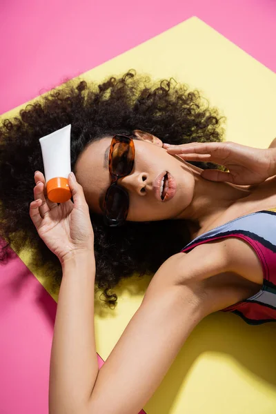 Кучерява афро-американська модель в модних сонцезахисних окулярах і смугастий купальник, що тримає сонцезахисний крем під час отримання засмаги на рожевому — стокове фото