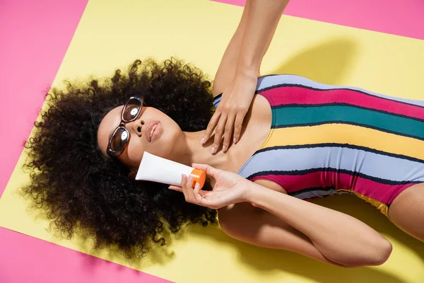 Top view of curly african american model in sunglasses and striped swimwear holding sunscreen while getting tan on pink — Stock Photo