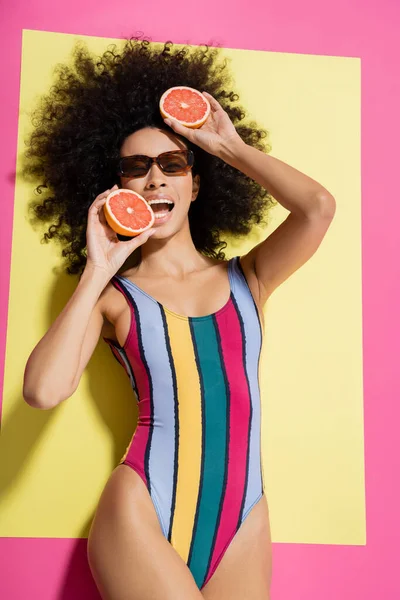 Сверху вид на кудрявую американку в солнечных очках и красочные купальники, кусающие грейпфрут на желтом и розовом фоне — стоковое фото