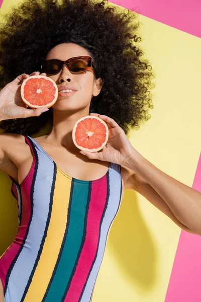 Top view of curly african american woman in sunglasses and colorful swimsuit holding juicy grapefruit halves on yellow background — Stock Photo