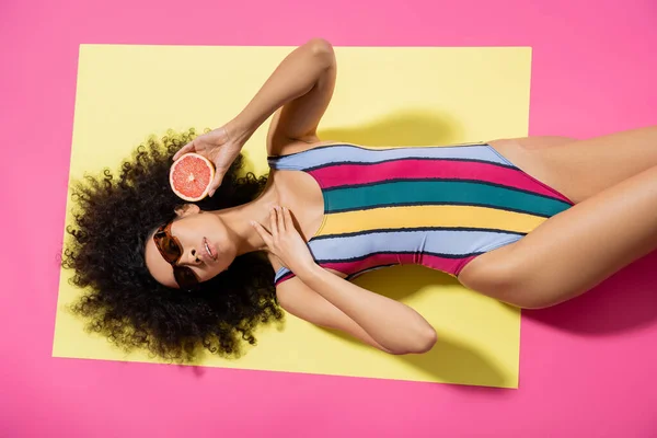 Вид зверху на молоду афроамериканку в сонцезахисних окулярах і барвистий купальник, що тримає грейпфрут наполовину на жовтому фоні — стокове фото