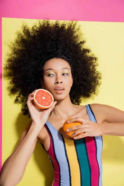 Top view of curly african american woman in colorful swimsuit holding grapefruit halves on yellow and pink background — Stock Photo
