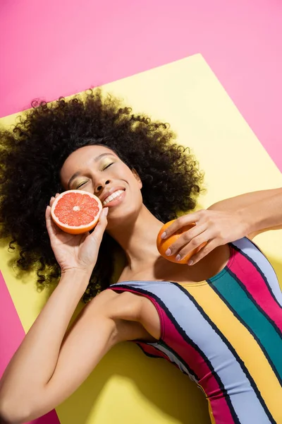 High angle view of pleased african american woman in colorful swimsuit holding grapefruit halves on yellow and pink — Stock Photo