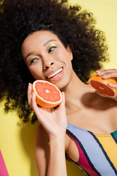 Top view of cheerful african american woman in colorful swimsuit holding grapefruit halves on yellow background — Stock Photo