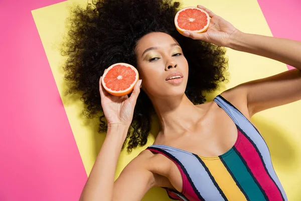Top view of curly african american model in colorful swimsuit holding grapefruit halves on yellow and pink background — Stock Photo