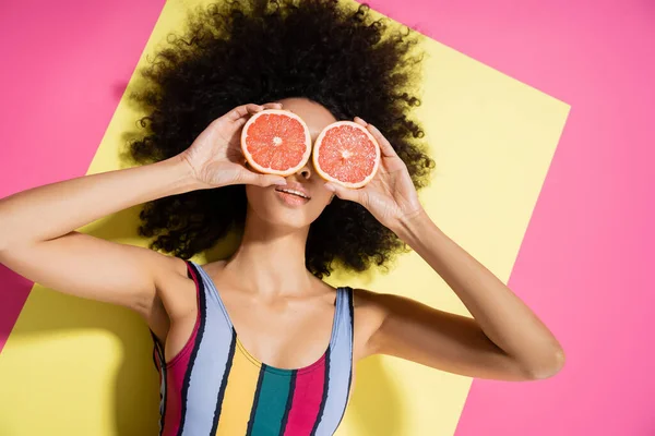Top view of curly african american woman in colorful swimsuit covering eyes with grapefruit halves on yellow and pink background — Stock Photo