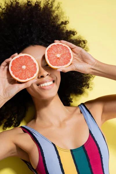 Top view of happy african american woman in colorful swimsuit covering eyes with grapefruit halves on yellow background — Stock Photo