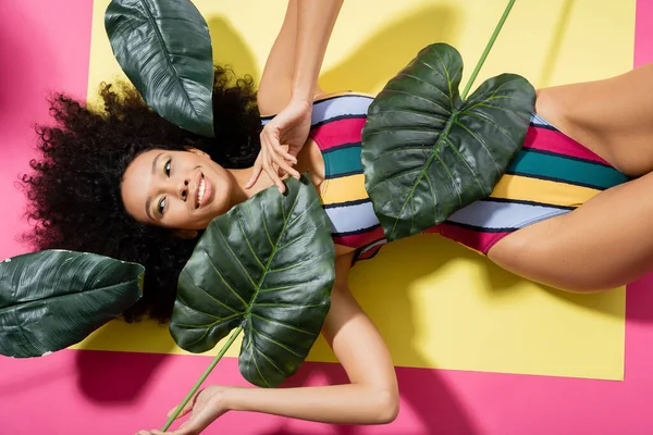 Draufsicht der fröhlichen afrikanisch-amerikanischen Frau im bunten Badeanzug, die sich in der Nähe grüner Palmblätter auf rosa bräunt — Stockfoto