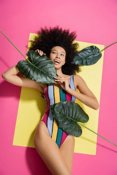 Vue de dessus de femme afro-américaine excitée en maillot de bain coloré obtenir bronzage près de feuilles de palmier vert sur rose — Photo de stock
