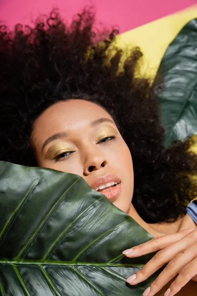 Vue du dessus de la femme afro-américaine bouclée bronzant près des feuilles de palmier vert sur rose — Photo de stock