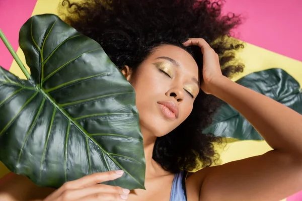 Vista superior de la mujer americana africana rizada con los ojos cerrados broncearse cerca de hojas de palma verde en rosa - foto de stock